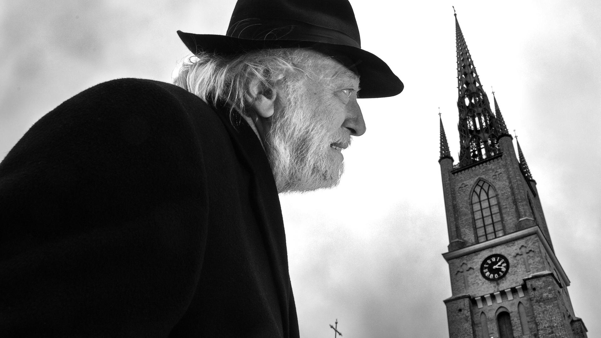 Autor László Krasznahorkai auf einem schwarz-weiß Bild in gebückter Haltung und mit Hut. Rechts daneben sieht man einen alten Kirchturm.
