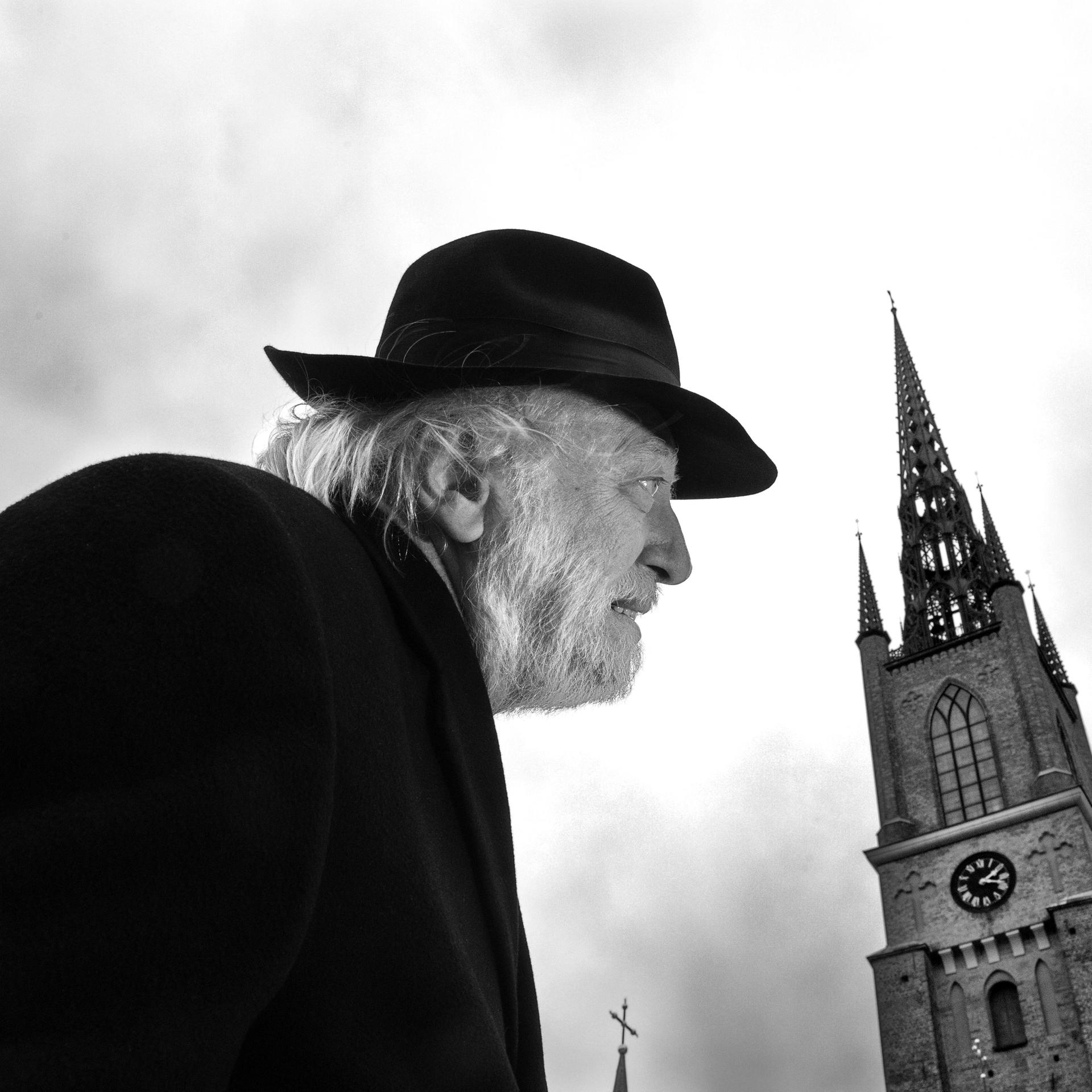 Autor László Krasznahorkai auf einem schwarz-weiß Bild in gebückter Haltung und mit Hut. Rechts daneben sieht man einen alten Kirchturm.