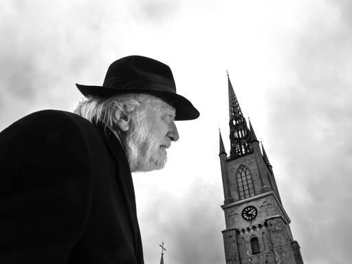 Autor László Krasznahorkai auf einem schwarz-weiß Bild in gebückter Haltung und mit Hut. Rechts daneben sieht man einen alten Kirchturm.