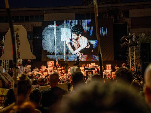 Es ist dunkel. In der unteren Bildhälfte sieht man beleuchtet eine große Menge Demonstranten mit Transparenten und Schildern. Sie schauen auf eine Leinwand. Dort läuft das Video von einer Geisel.