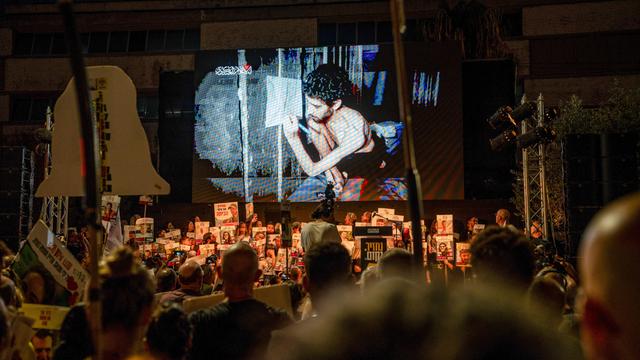 Es ist dunkel. In der unteren Bildhälfte sieht man beleuchtet eine große Menge Demonstranten mit Transparenten und Schildern. Sie schauen auf eine Leinwand. Dort läuft das Video von einer Geisel.