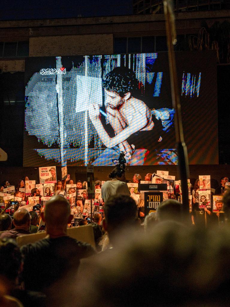 Es ist dunkel. In der unteren Bildhälfte sieht man beleuchtet eine große Menge Demonstranten mit Transparenten und Schildern. Sie schauen auf eine Leinwand. Dort läuft das Video von einer Geisel.