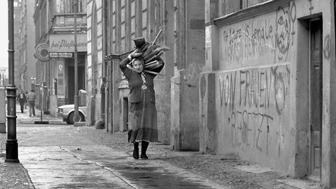 Eine Frau mit Kopftuch trägt einen Stapel Holzlatten auf der Schulter, an den Häuserwänden sind Graffiti zu sehen.