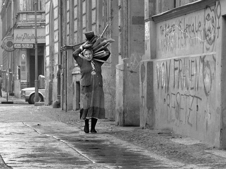Eine Frau mit Kopftuch trägt einen Stapel Holzlatten auf der Schulter, an den Häuserwänden sind Graffiti zu sehen.