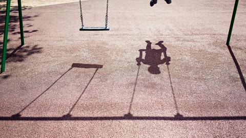Der Schatten eines Kindes auf einer Schaukel ist auf dem Betonboden eines Spielplatzes zu sehen.