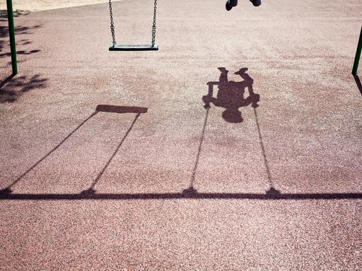 Der Schatten eines Kindes auf einer Schaukel ist auf dem Betonboden eines Spielplatzes zu sehen.