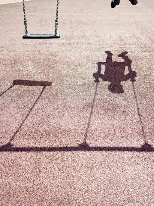 Der Schatten eines Kindes auf einer Schaukel ist auf dem Betonboden eines Spielplatzes zu sehen.