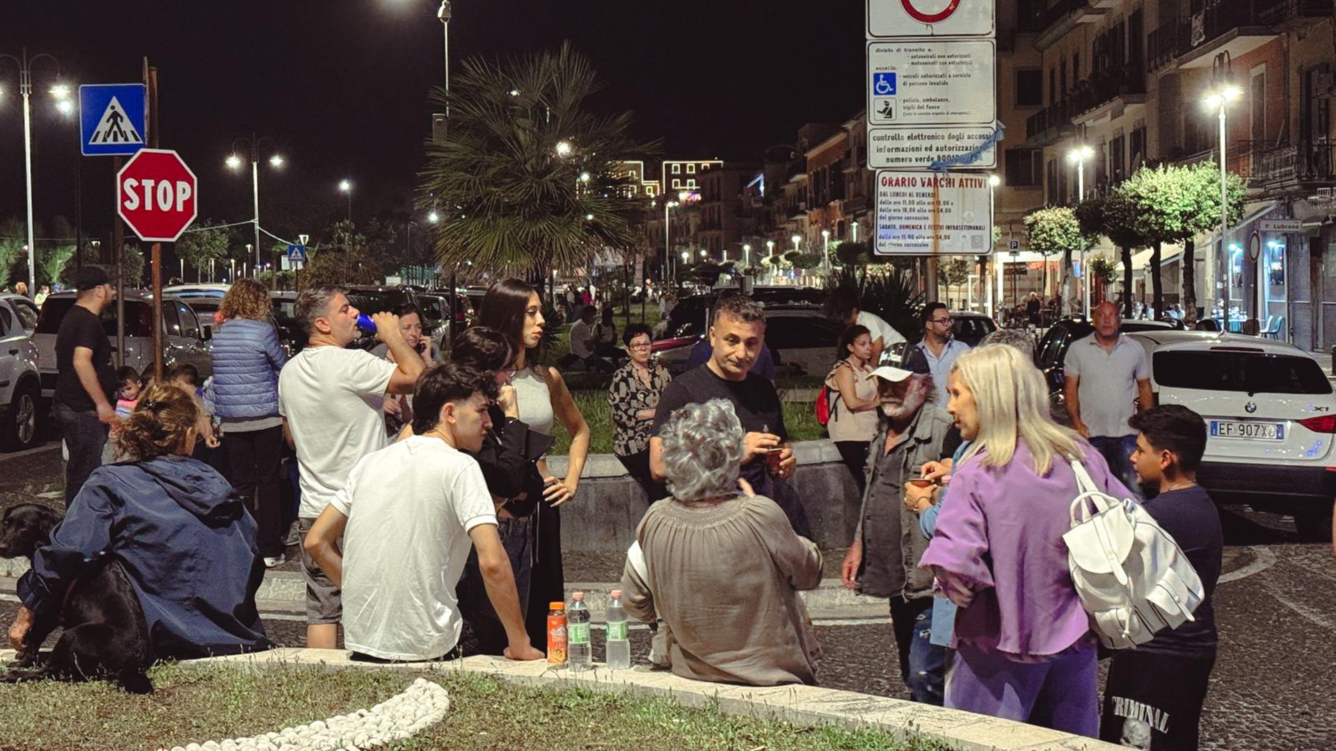 Es ist Nacht. Viele Menschen stehen auf einer Straße an begrünten Anlagen und reden miteinander. Sie werden von Straßenlaternen beleuchtet. Es ist Nacht. Viele Menschen stehen auf einer Straße an begrünten Anlagen und reden miteinander. Sie werden von Straßenlaternen beleuchtet.