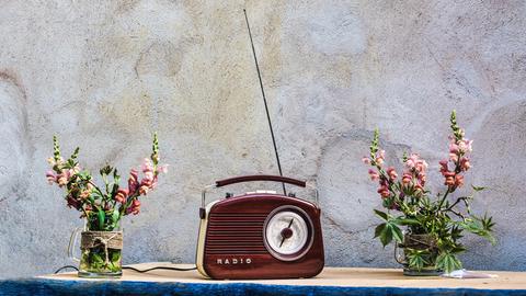 Auch wenn Radios längst nicht mehr so aussehen, fangen die Macher von „Angel Radio“ immer noch den Sound von Damals ein. Zu sehen: Ein rotbraunes, altes Handkofferradio mit ausgezogener Antenne in der Mitte eines Tisches. Daneben jeweils eine Blumenvase mit Löwenmäulchen. 