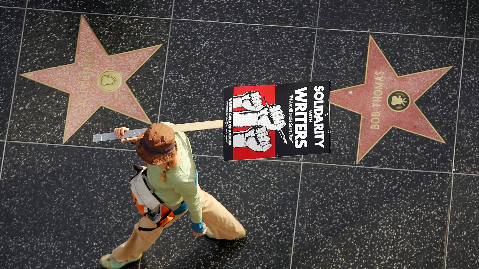 Aufnahme aus der Vogelperspektive zeigt eine Gewerkschaftsmitglied mit einem Plakat auf dem steht "Solidarity with Writers" zwischen zwei Sternen auf  dem Hollywood Walk of Fame.