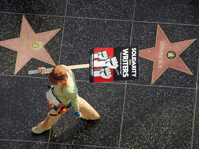 Aufnahme aus der Vogelperspektive zeigt eine Gewerkschaftsmitglied mit einem Plakat auf dem steht "Solidarity with Writers" zwischen zwei Sternen auf  dem Hollywood Walk of Fame.