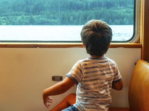 Ein Junge sitzt an einem Zugfenster und schaut auf das Wasser hinaus. Der Junge trägt ein gestreiftes Hemd.