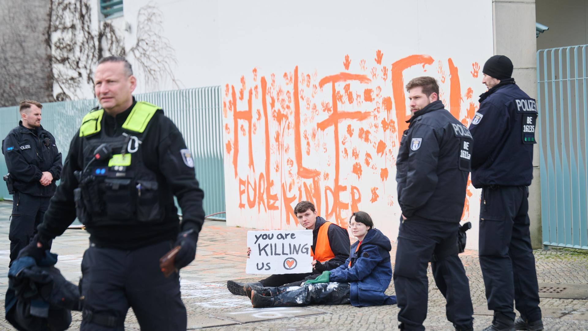 "Hilfe! Eure Kinder" steht mit der Farbe Orange ans Kanzleramt geschrieben. Klimaaktivisten der Letzten Generation sitzen davor. Sie halten ein Schild in der Hand. Darauf steht: "You are kliing us" - Ihr tötet uns. Außerdem sind vier Polizeibeamte zu sehen. "Hilfe! Eure Kinder" steht mit der Farbe Orange ans Kanzleramt geschrieben. Klimaaktivisten der Letzten Generation sitzen davor. Sie halten ein Schild in der Hand. Darauf steht: "You are kliing us" - Ihr tötet uns. Außerdem sind vier Polizeibeamte zu sehen.
