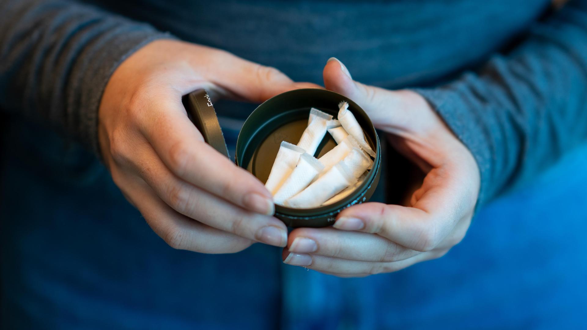 Blick von oben auf die Hände einer Frau, die eine Dose mit weißen, kleinen Päckchen hält, die man Pouches, Nicopods, Zyn oder Snus nennt.