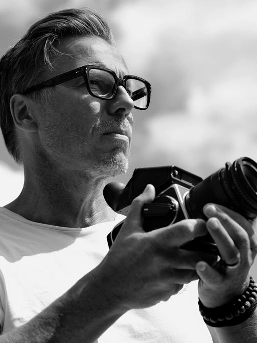 Schwarzweißporträt von Olaf Heine. Ein Mann mit Dreitagebart und schwarzer Brille, der eine Mittelformatkamera in der Hand hält.