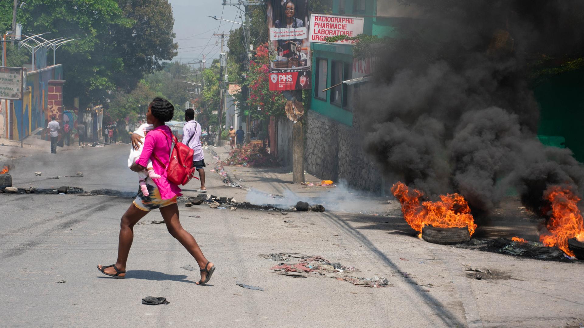 Eine Frau mit einem Kind im Arm rennt über eine Straße in Haitis Hauptstadt Port-au-Prince. Neben ihr sind brennende Reifen zu sehen, auf der Straße liegt Müll.