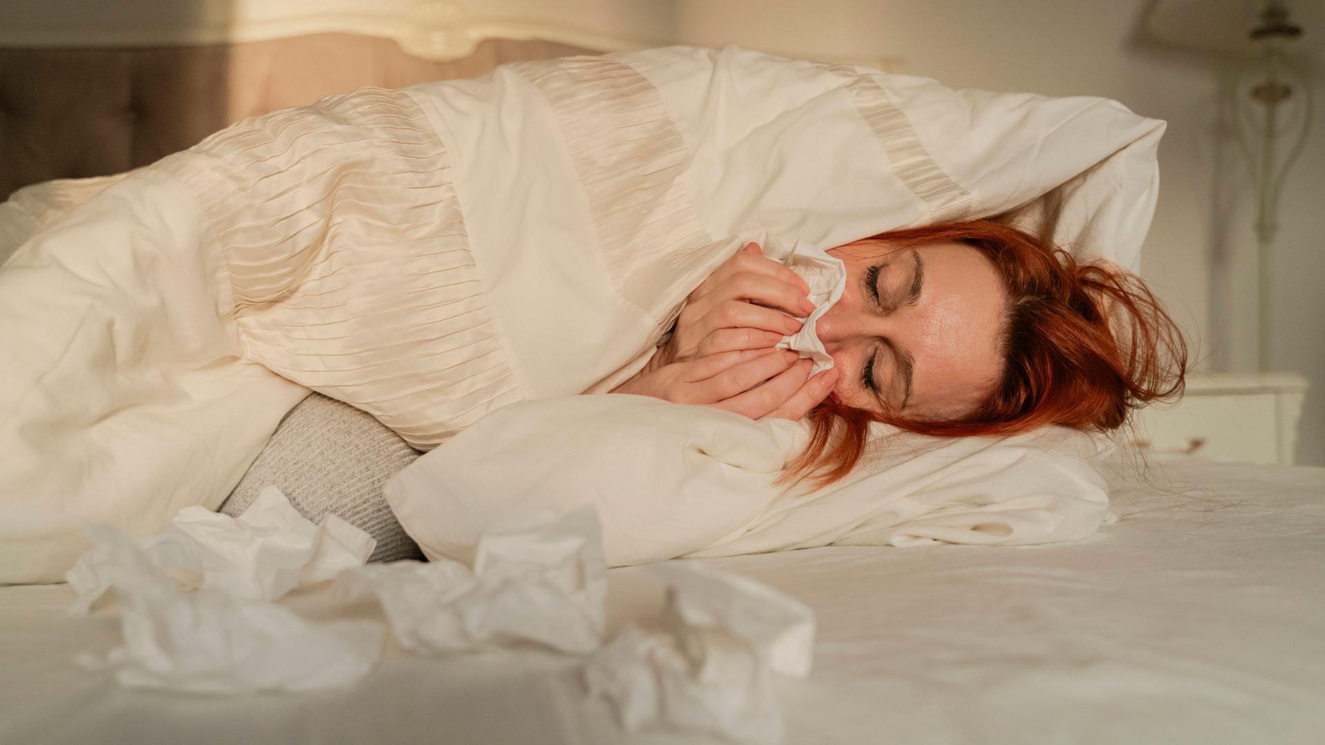 Eine Frau mit roten Haaren liegt zugedeckt in einem Bett und putzt sich die Nase mit einem Taschentuch. Vor ihr im Bett liegen mehrere bentutzte Taschentücher.