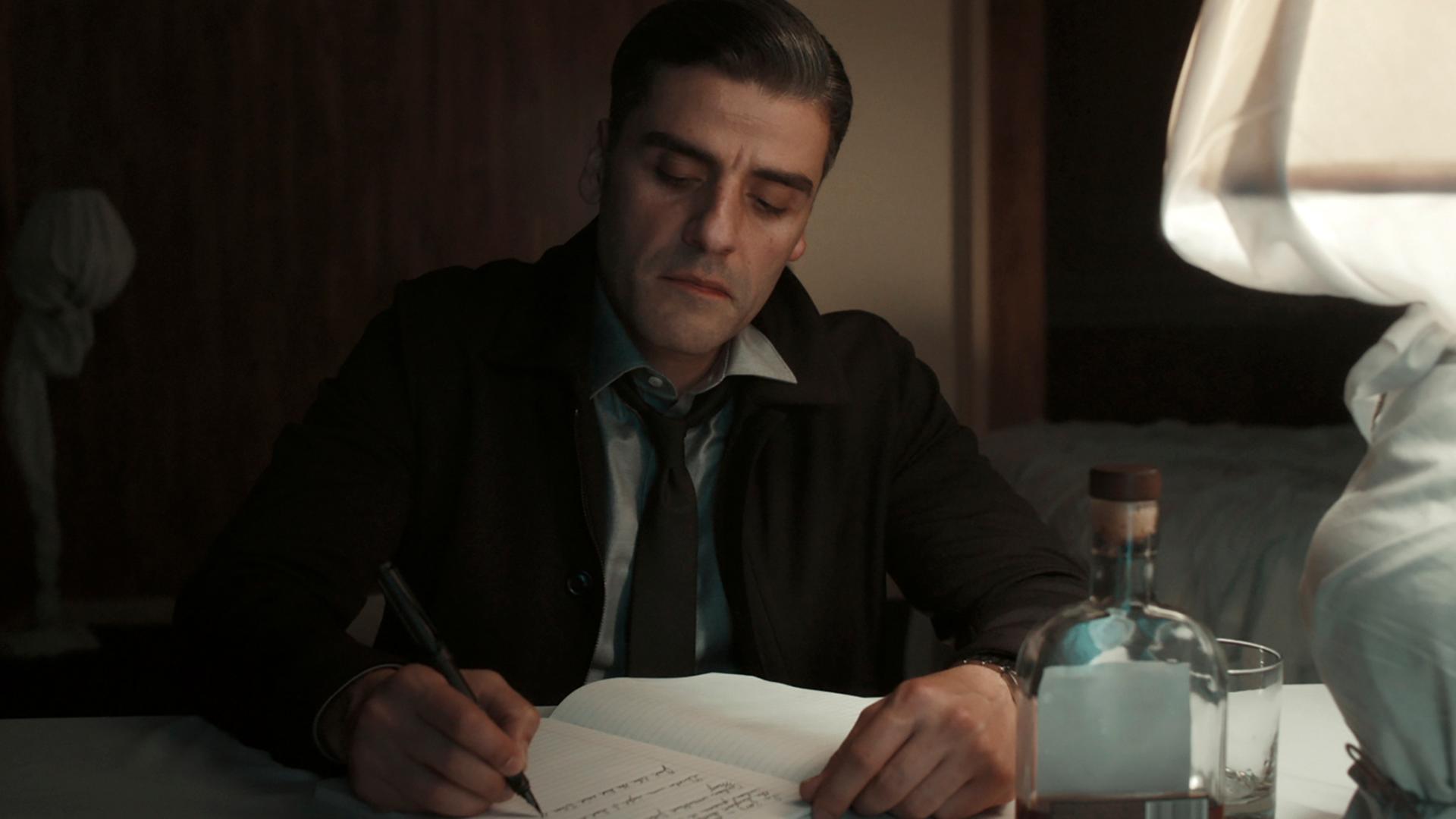Oscar Isaac als William Tell in einer Szene aus "The Card Counter": Er sitzt an einem Schreibtisch in einem abgedunkelten Hotelzimmer und schreibt in sein Tagebuch.