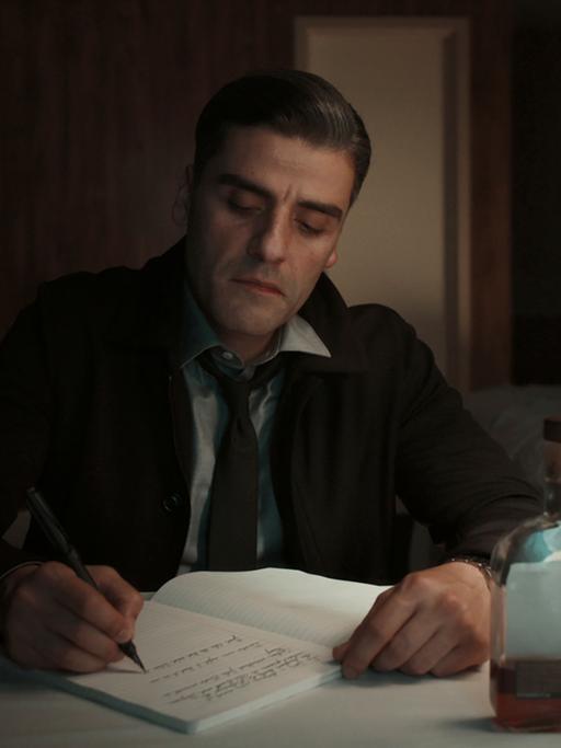 Oscar Isaac als William Tell in einer Szene aus "The Card Counter": Er sitzt an einem Schreibtisch in einem abgedunkelten Hotelzimmer und schreibt in sein Tagebuch.