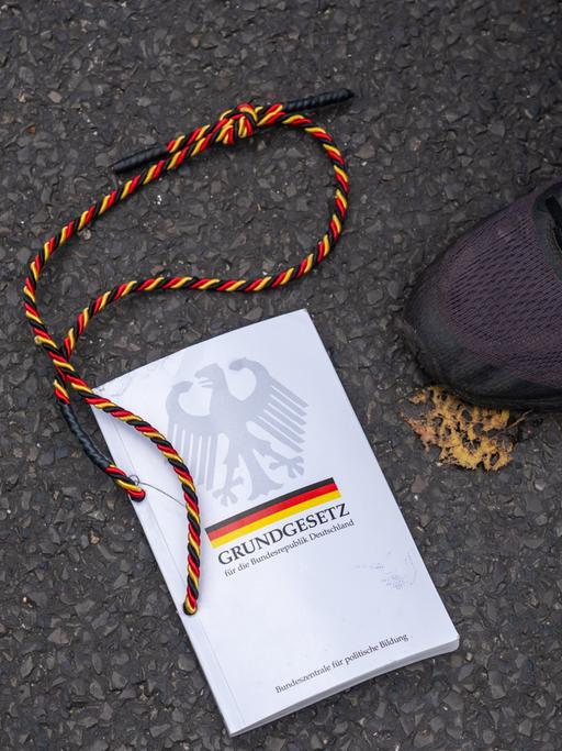 Auf der Straße liegt eine Ausgabe des Grundgesetzes. Daneben ist ein Schuh. An dem Buch ist ein schwarz-rot-gelbes Band angebracht.