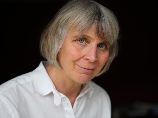 Porträt der Philosophin Barbara Schmitz. Sie trägt kutze, graue Haare und eine weiße Bluse vor schwarzem Hintergrund. 