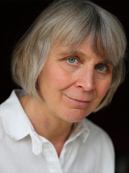 Porträt der Philosophin Barbara Schmitz. Sie trägt kutze, graue Haare und eine weiße Bluse vor schwarzem Hintergrund. 