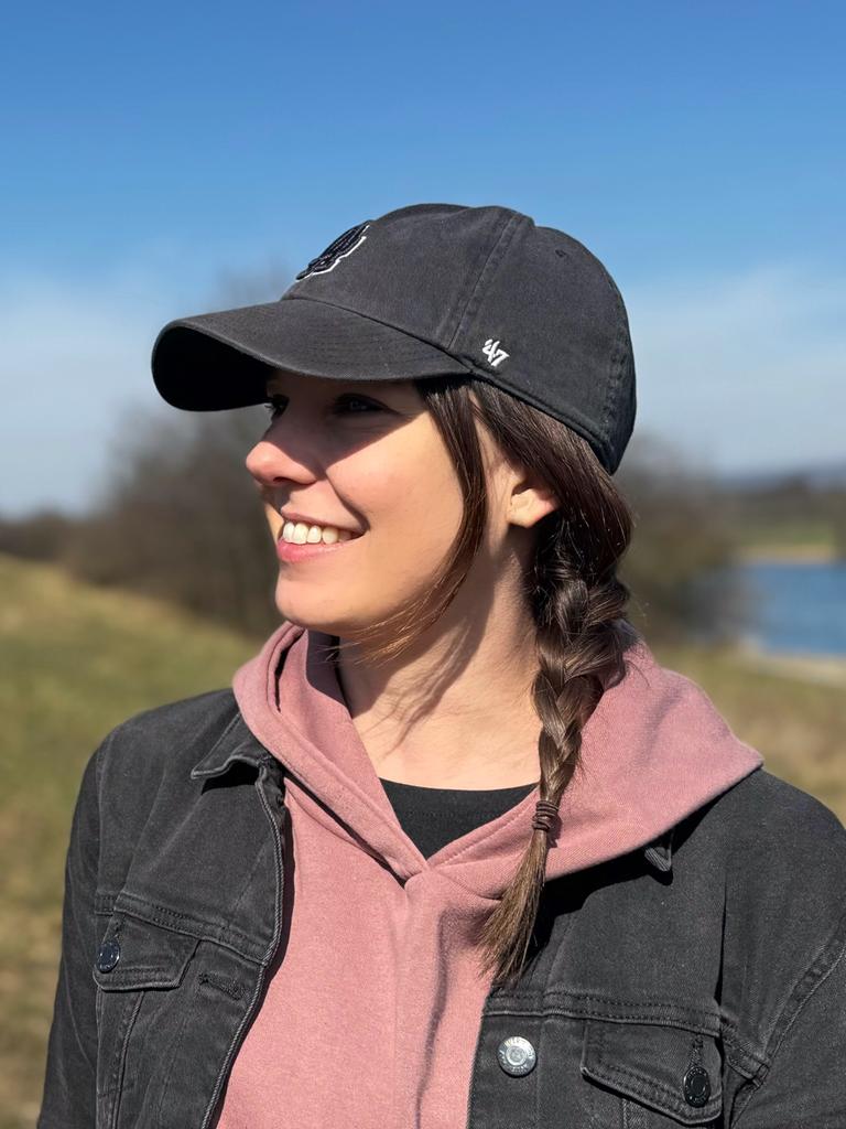 Porträt der Protagonistin Conny, die einen geflochtenen Zopf unter ihrem schwarzen Basecap trägt. Sie steht auf einer Wiese am Ufer eines Flusses.