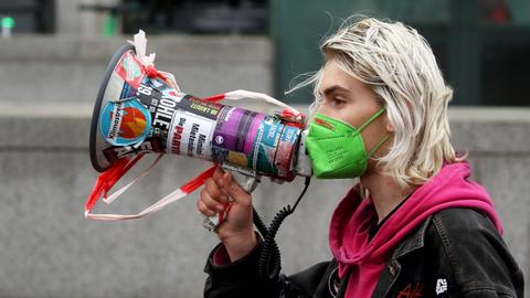 Eine junge Frau mit grünem Mundschutz hält bei einer Demonstration ein bunt beklebtes Megafon vor ihr Gesicht. Eine junge Frau mit grünem Mundschutz hält bei einer Demonstration ein bunt beklebtes Megafon vor ihr Gesicht.