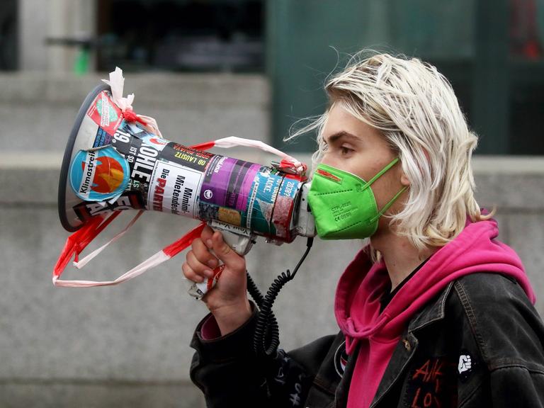 Eine junge Frau mit grünem Mundschutz hält bei einer Demonstration ein bunt beklebtes Megafon vor ihr Gesicht.