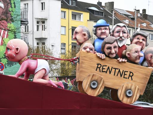 Rosenmontagszug in Düsseldorf: ein Themenwagen zeigt ein Baby, das mühevoll einen Karren mit Rentnern zieht.