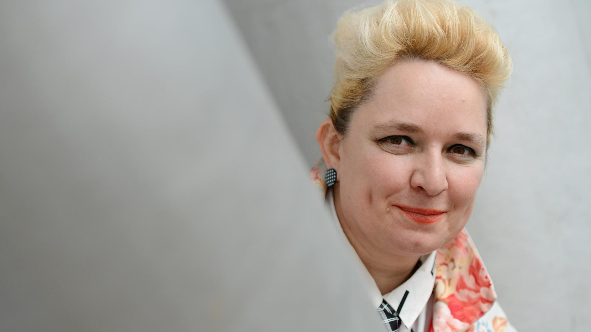 Eine Frau mit blondierten Haaren schaut um einen Vorsprung und lächelt in die Kamera. Es ist die Künstlerin Mariola Brillowska. Eine Frau mit blondierten Haaren schaut um einen Vorsprung und lächelt in die Kamera. Es ist die Künstlerin Mariola Brillowska.