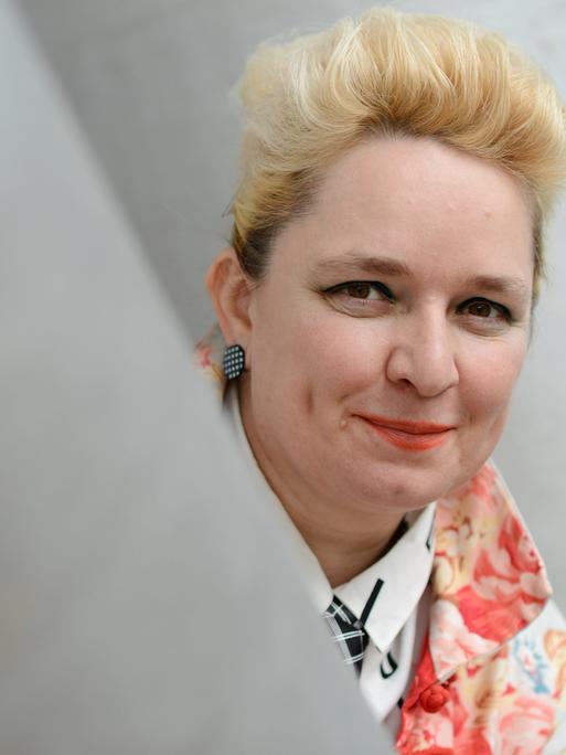 Eine Frau mit blondierten Haaren schaut um einen Vorsprung und lächelt in die Kamera. Es ist die Künstlerin Mariola Brillowska.