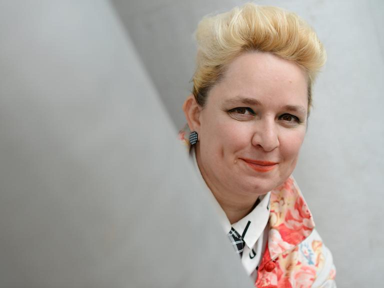 Eine Frau mit blondierten Haaren schaut um einen Vorsprung und lächelt in die Kamera. Es ist die Künstlerin Mariola Brillowska.