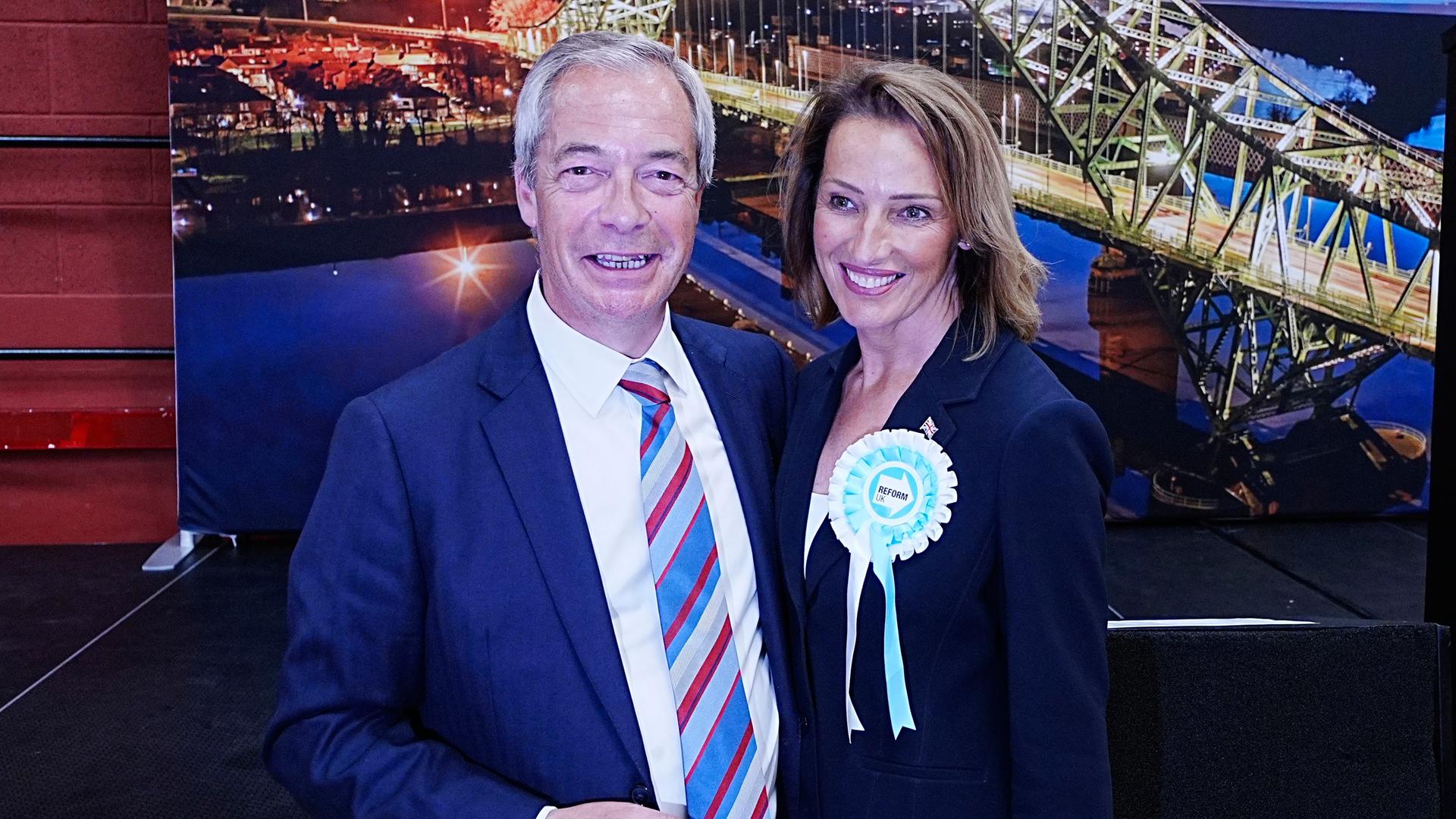 Reform UK-Chef Nigel Farage und die Kandidatin Sarah Pochin stehen nebeneinander, halten sich im Arm und lächeln. 