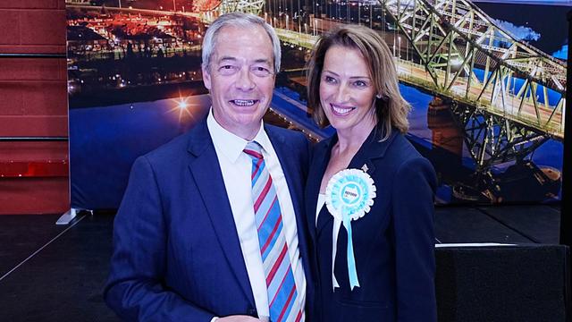 Reform UK-Chef Nigel Farage und die Kandidatin Sarah Pochin stehen nebeneinander, halten sich im Arm und lächeln. 