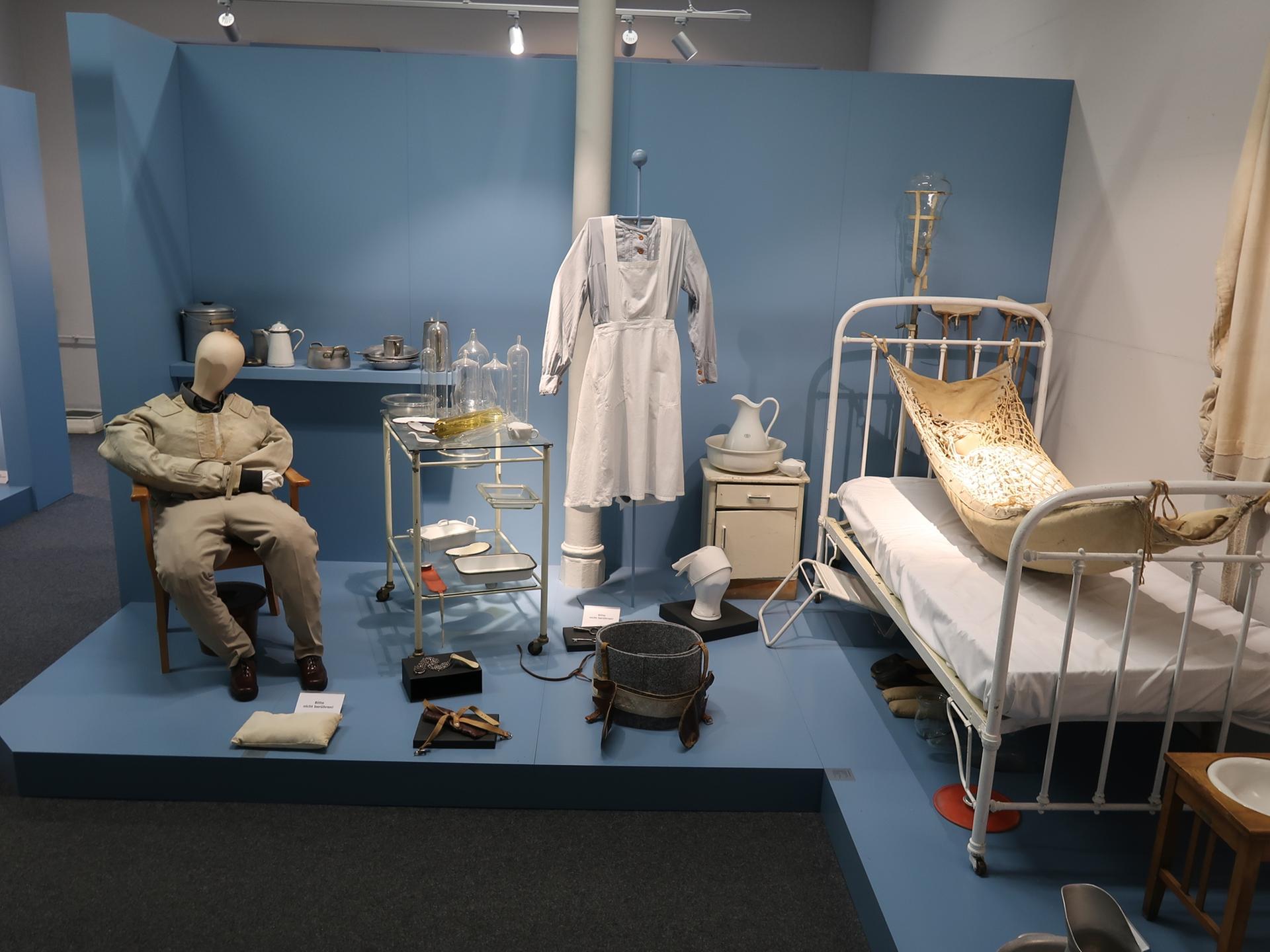 Ausstellungsraum: Ein Krankenzimmer aus dem vorigen Jahrhundert, man sieht ein krankenbett und medizinische Utensilien, eine Schwesternuniform und eine Puppe in Erwachsenengröße, die auf einem Stuhl sitzt