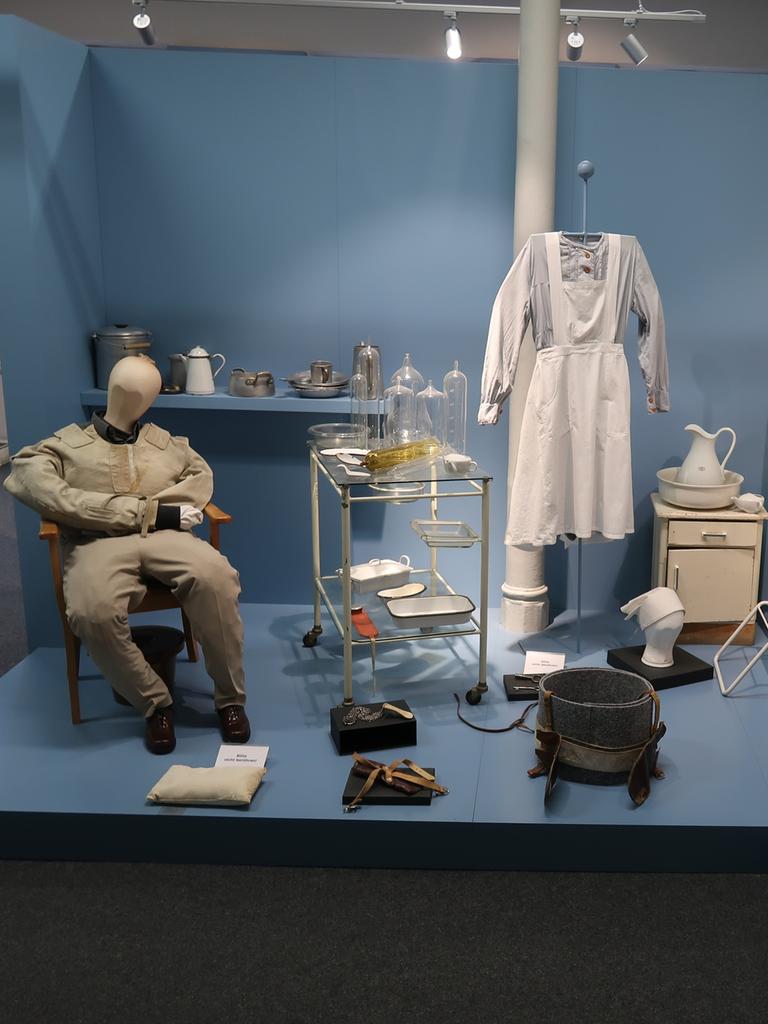 Ausstellungsraum: Ein Krankenzimmer aus dem vorigen Jahrhundert, man sieht ein krankenbett und medizinische Utensilien, eine Schwesternuniform und eine Puppe in Erwachsenengröße, die auf einem Stuhl sitzt