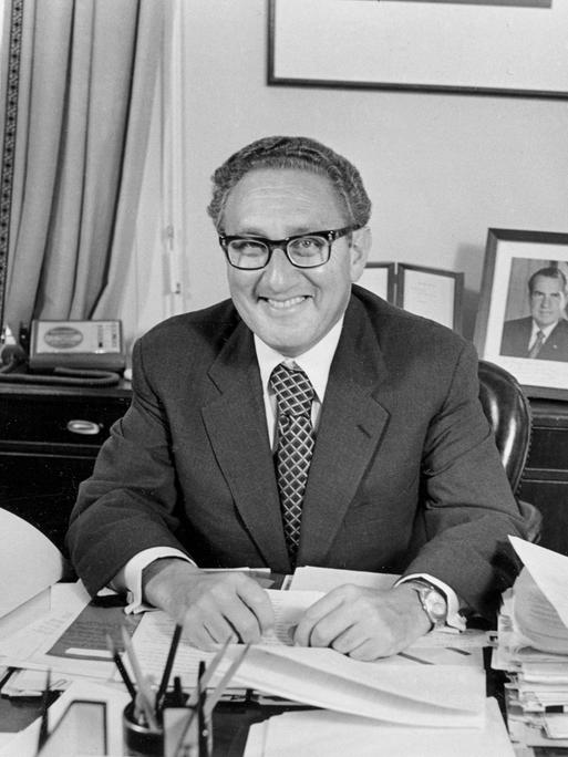 Henry Kissinger sitzt an einem Schreibtisch und lacht