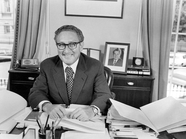 Henry Kissinger sitzt an einem Schreibtisch und lacht