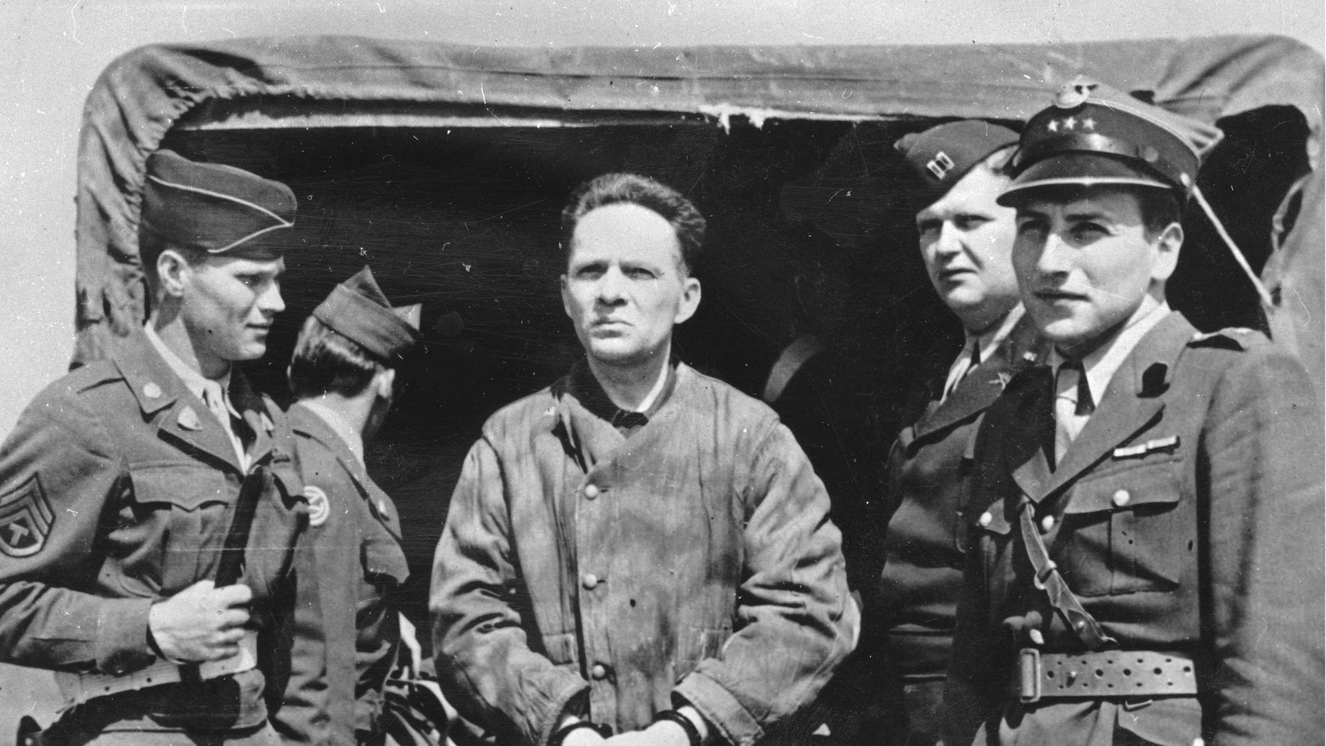Der Kommandant des KZ Auschwitz Rudolf Höß 1946 auf dem Nürnberger Flugplatz auf dem Weg zu den Nürnberger Prozessen. Vier alliierte Soldaten stehen auf dem Schwarzweißbild um ihn herum und bewachen ihn.