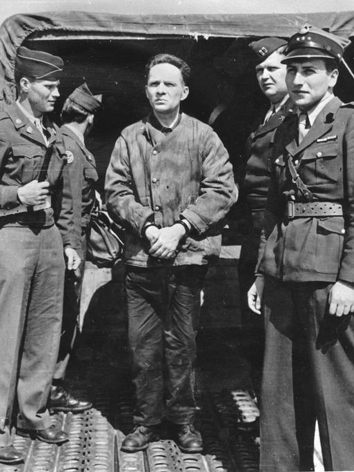 Der Kommandant des KZ Auschwitz Rudolf Höß 1946 auf dem Nürnberger Flugplatz auf dem Weg zu den Nürnberger Prozessen. Vier alliierte Soldaten stehen auf dem Schwarzweißbild um ihn herum und bewachen ihn.