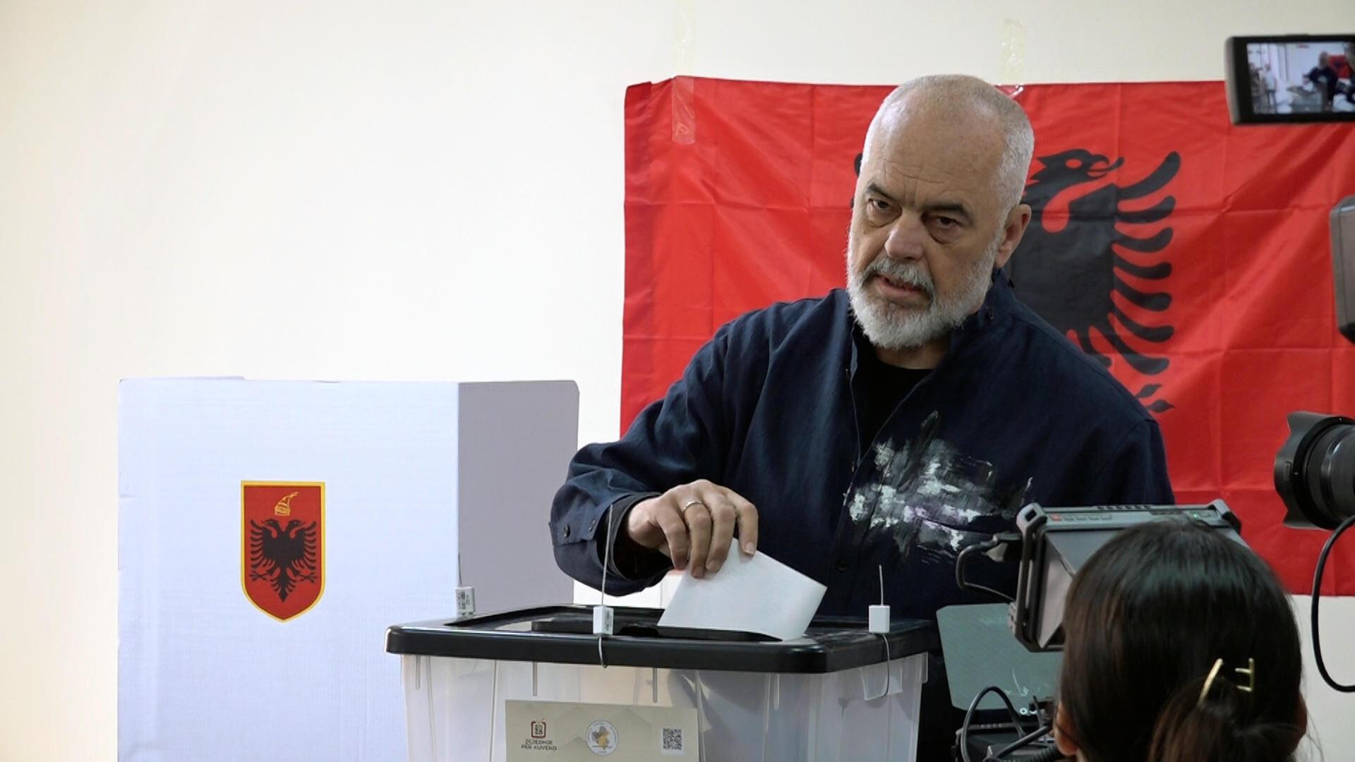 Ein Mann mit grauen Haaren und Vollbart wirft einen Stimmzettel in eine Wahlurne. Im Hintergrund ist eine rote albanische Flagge zu sehen, im Vordergrund ist eine Fernsehkamera. Ein Mann mit grauen Haaren und Vollbart wirft einen Stimmzettel in eine Wahlurne. Im Hintergrund ist eine rote albanische Flagge zu sehen, im Vordergrund ist eine Fernsehkamera.