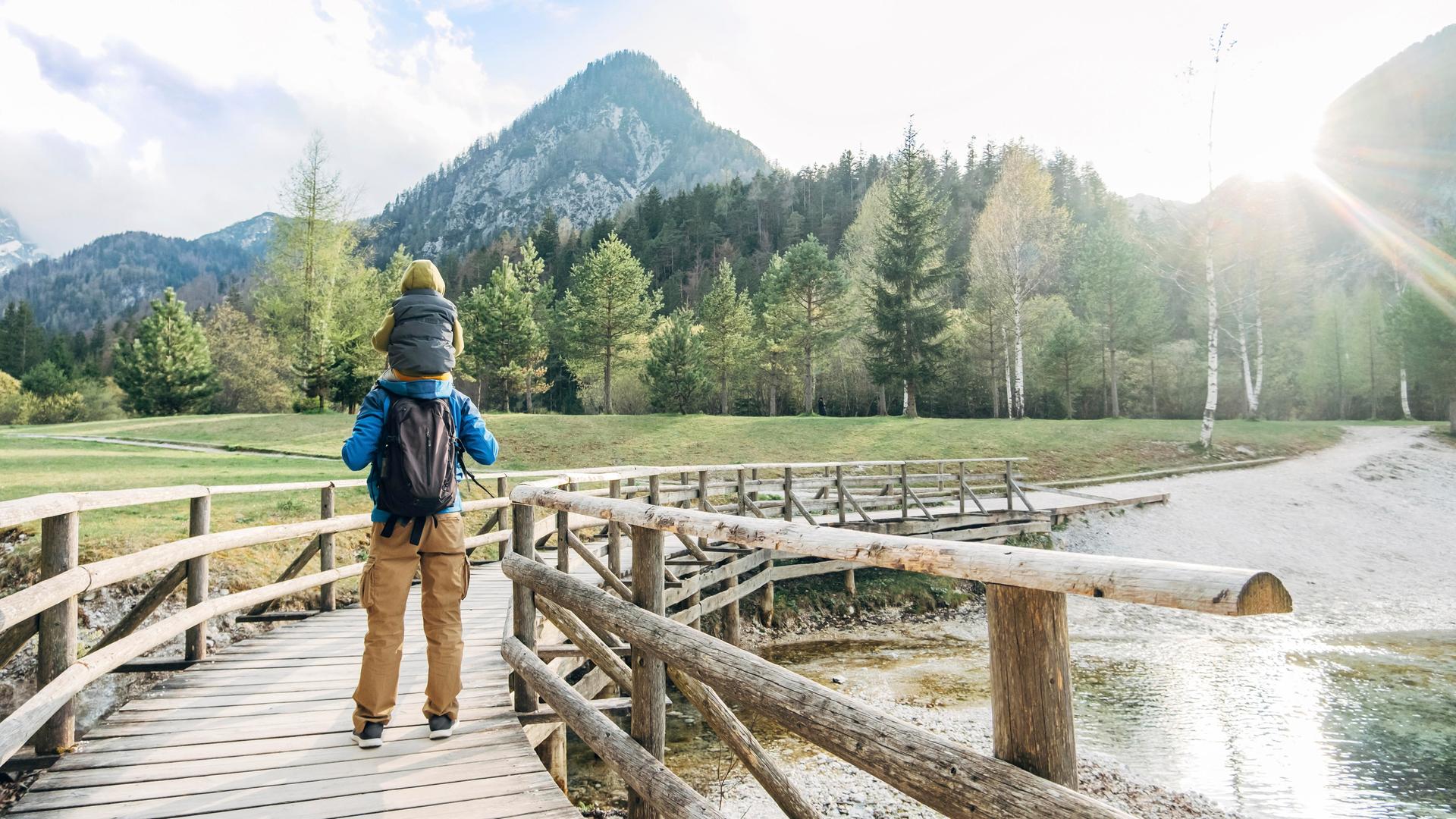 Ein Mann trägt ein Kind auf den Schultern und wandert über eine Holzbrücke in Richtung Berge Ein Mann trägt ein Kind auf den Schultern und wandert über eine Holzbrücke in Richtung Berge