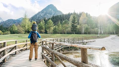 Ein Mann trägt ein Kind auf den Schultern und wandert über eine Holzbrücke in Richtung Berge