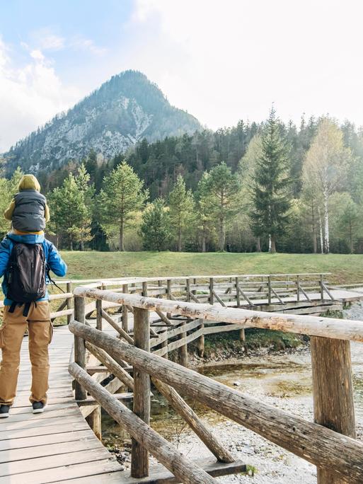 Ein Mann trägt ein Kind auf den Schultern und wandert über eine Holzbrücke in Richtung Berge