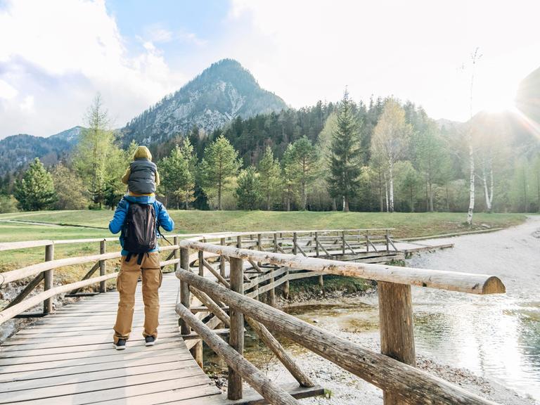 Ein Mann trägt ein Kind auf den Schultern und wandert über eine Holzbrücke in Richtung Berge