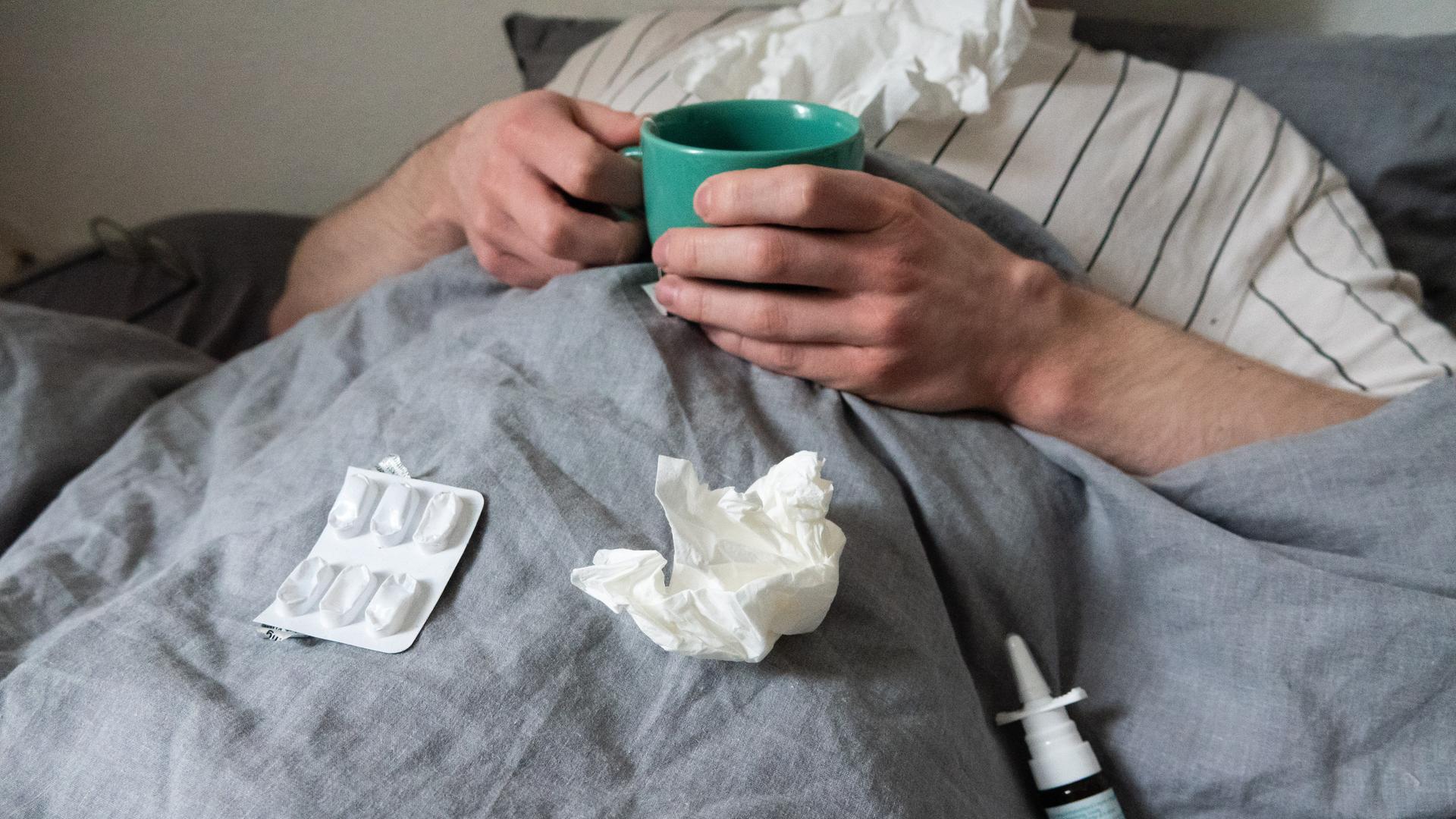 Ein Mann liegt mit Taschentüchern, Teetasse, Nasenspray und Tabletten im Bett unter einer grauen Decke. Sein Kopf ist nicht zu sehen.  (gestellte Szene). 