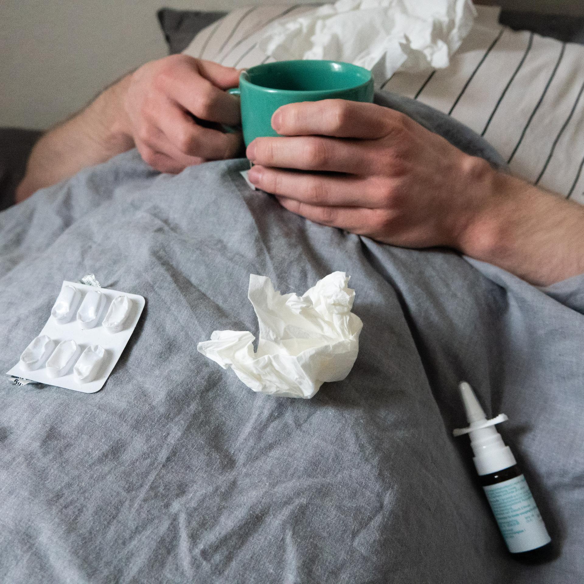 Ein Mann liegt mit Taschentüchern, Teetasse, Nasenspray und Tabletten im Bett unter einer grauen Decke. Sein Kopf ist nicht zu sehen.  (gestellte Szene). 