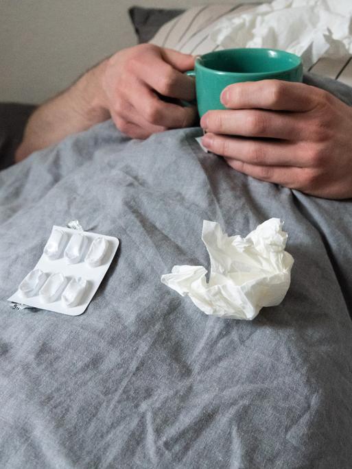 Ein Mann liegt mit Taschentüchern, Teetasse, Nasenspray und Tabletten im Bett unter einer grauen Decke. Sein Kopf ist nicht zu sehen.  (gestellte Szene). 