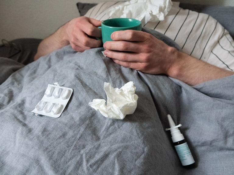 Ein Mann liegt mit Taschentüchern, Teetasse, Nasenspray und Tabletten im Bett unter einer grauen Decke. Sein Kopf ist nicht zu sehen.  (gestellte Szene). 